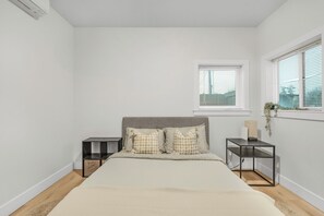 A bedroom with a bed, two bedside tables, a window with blinds, and a small plant on the window sill.