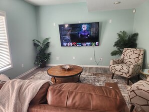 Uma sala de estar com televisão na parede, um sofá de couro marrom, uma poltrona estampada e uma mesinha com uma tigela.