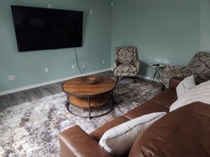 Un salon doté d’un grand téléviseur à écran plat fixé au mur, d’une table basse ronde en bois, d’un fauteuil à motifs et d’un canapé en cuir marron.