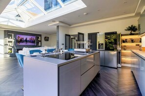 A modern kitchen with a central island, a dining area with a sofa, and a large TV screen. - 4 Bedroom Luxury Home Features Hottub & Pool Table (Northwood)