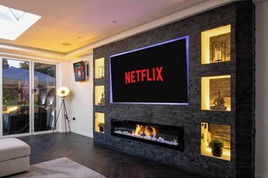 A modern living room with a large flat-screen TV mounted on a stone wall, a fireplace, and a glass door leading to an outdoor area. - 4 Bedroom Luxury Home Features Hottub & Pool Table (Northwood)