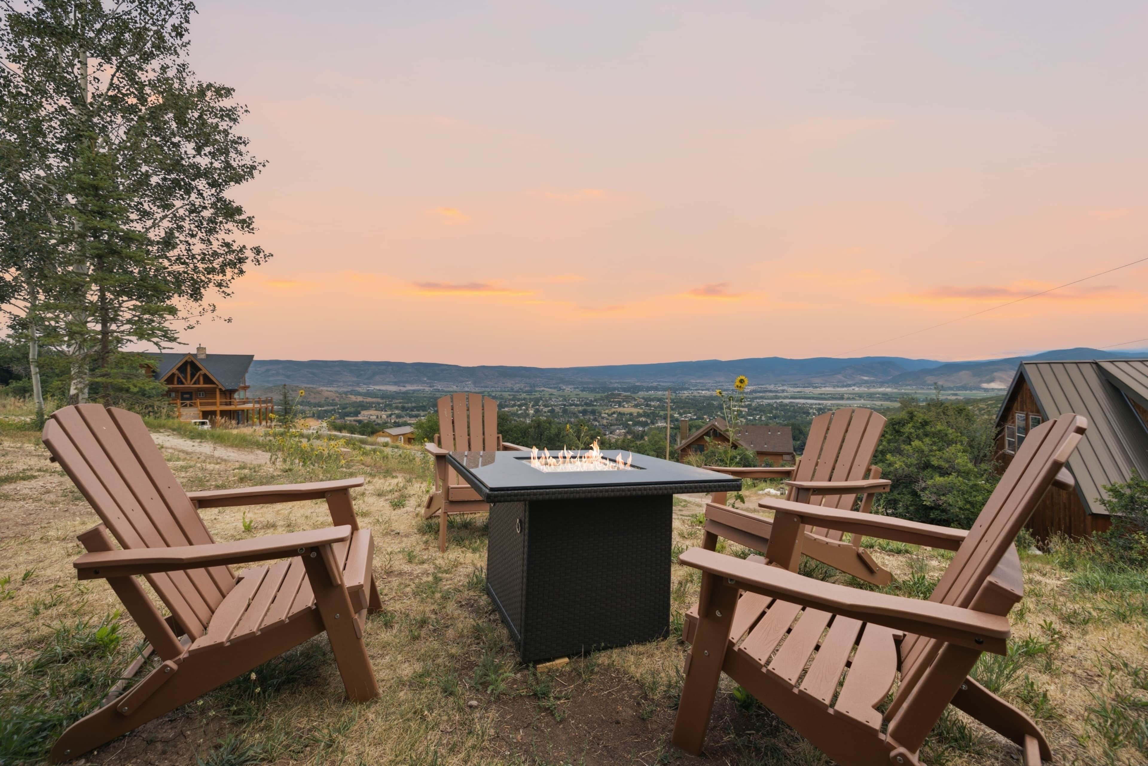 A wooden deck with Adirondack chairs and a fire pit, overlooking a scenic landscape with hills and a sunset.