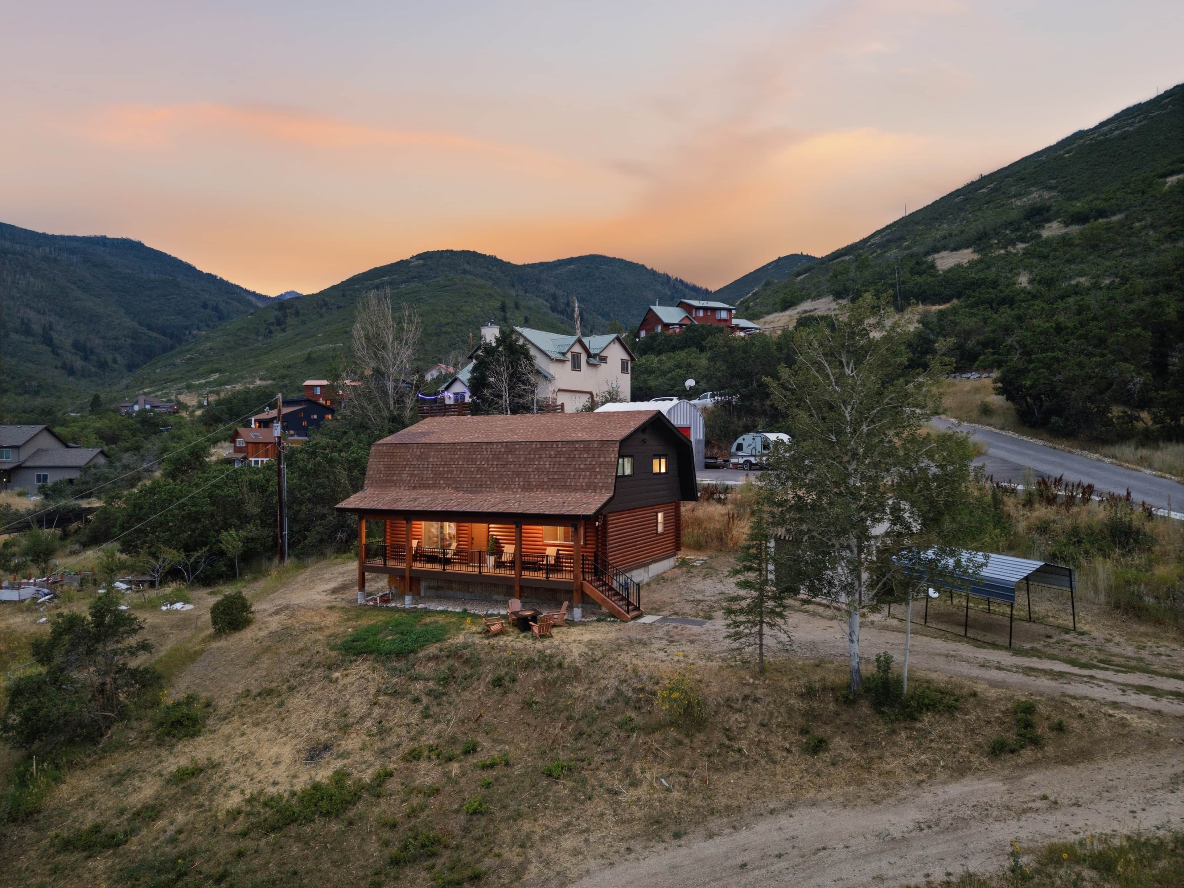 A cabin with a porch and outdoor seating, surrounded by a hilly landscape with houses in the background.