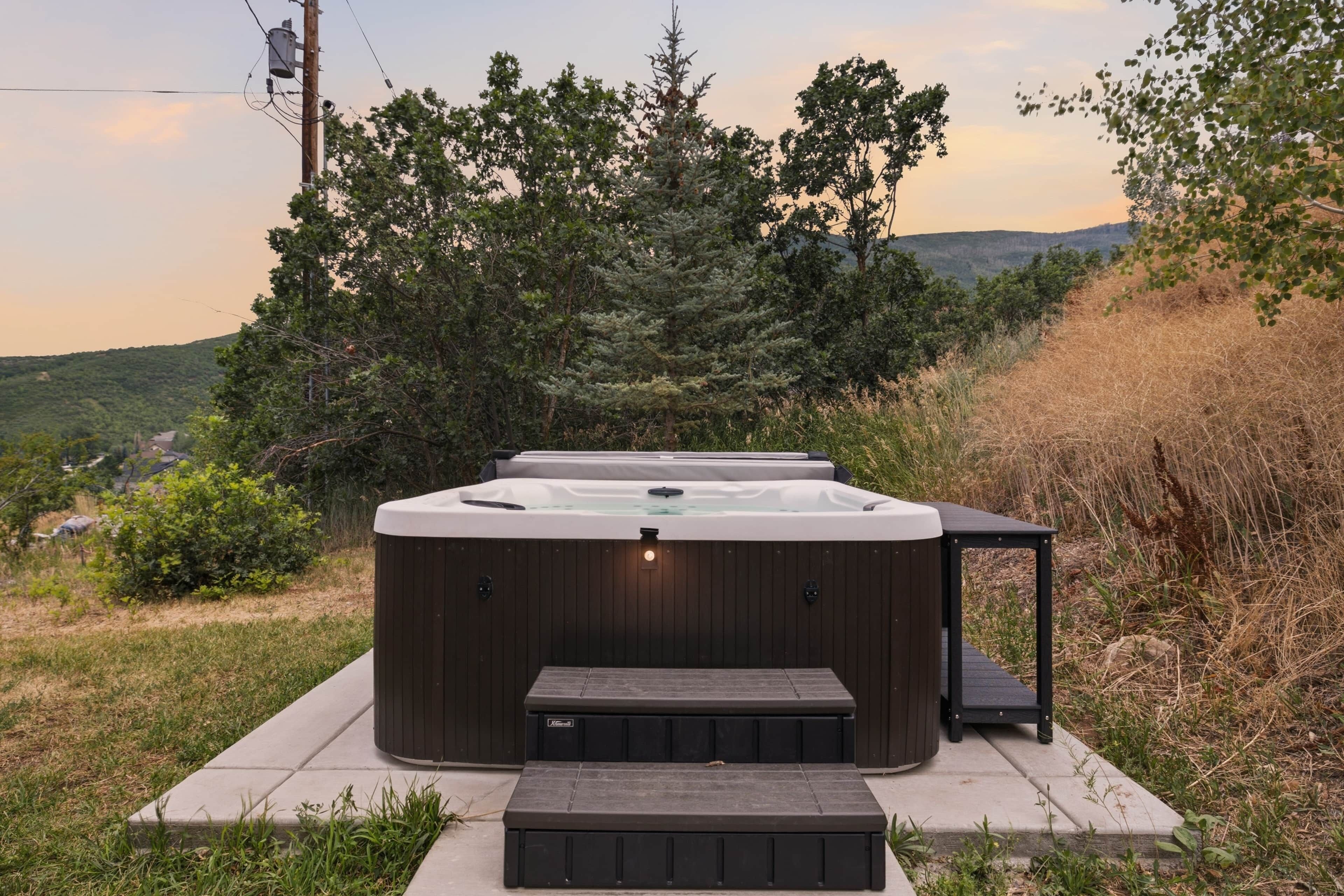 A hot tub with a wooden platform and steps, surrounded by greenery and hills.