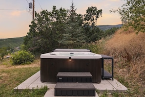 A hot tub with a wooden platform and steps, surrounded by greenery and hills.