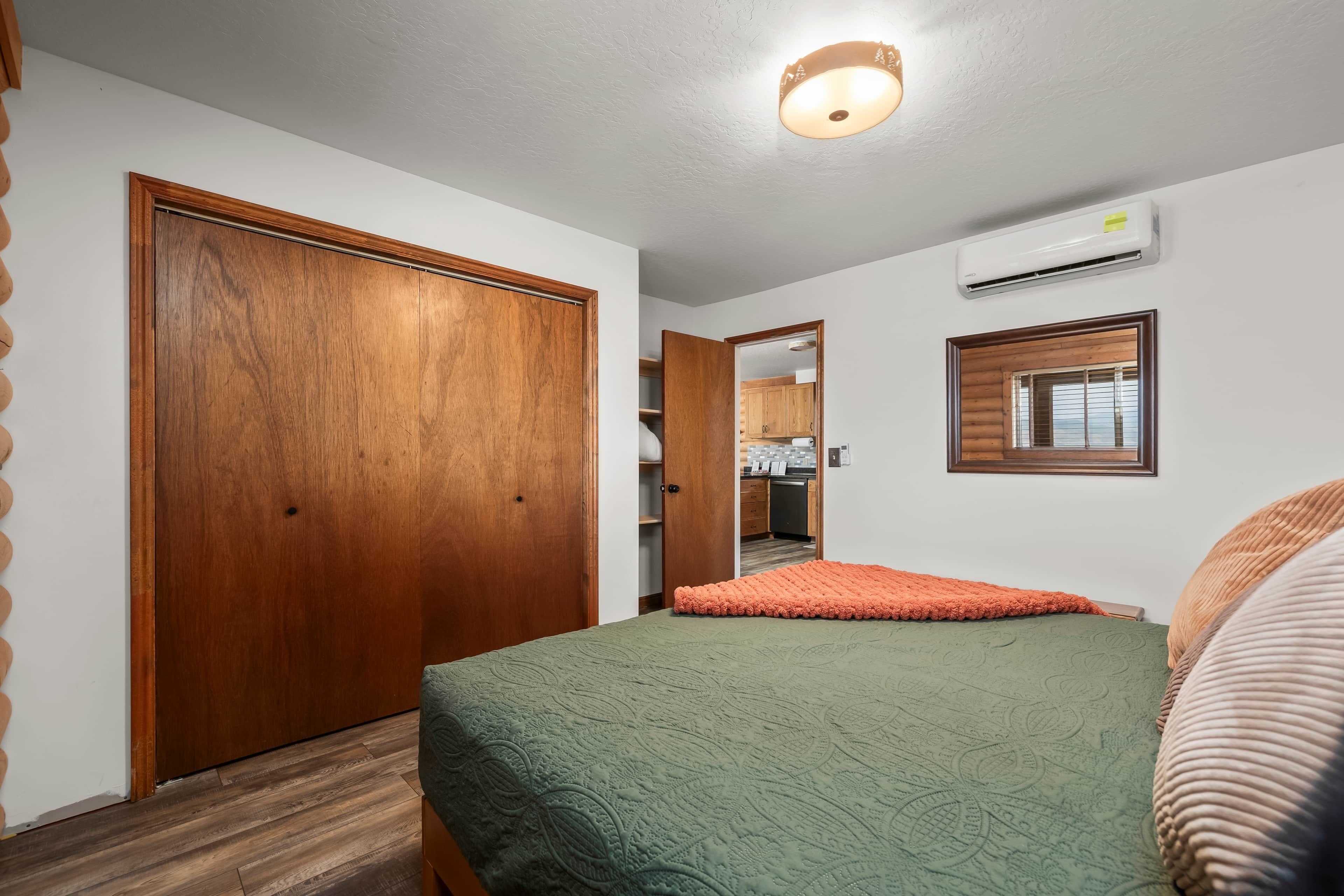A bedroom with a wooden wardrobe, a bed with a green quilt, a window with blinds, and an air conditioning unit on the wall.