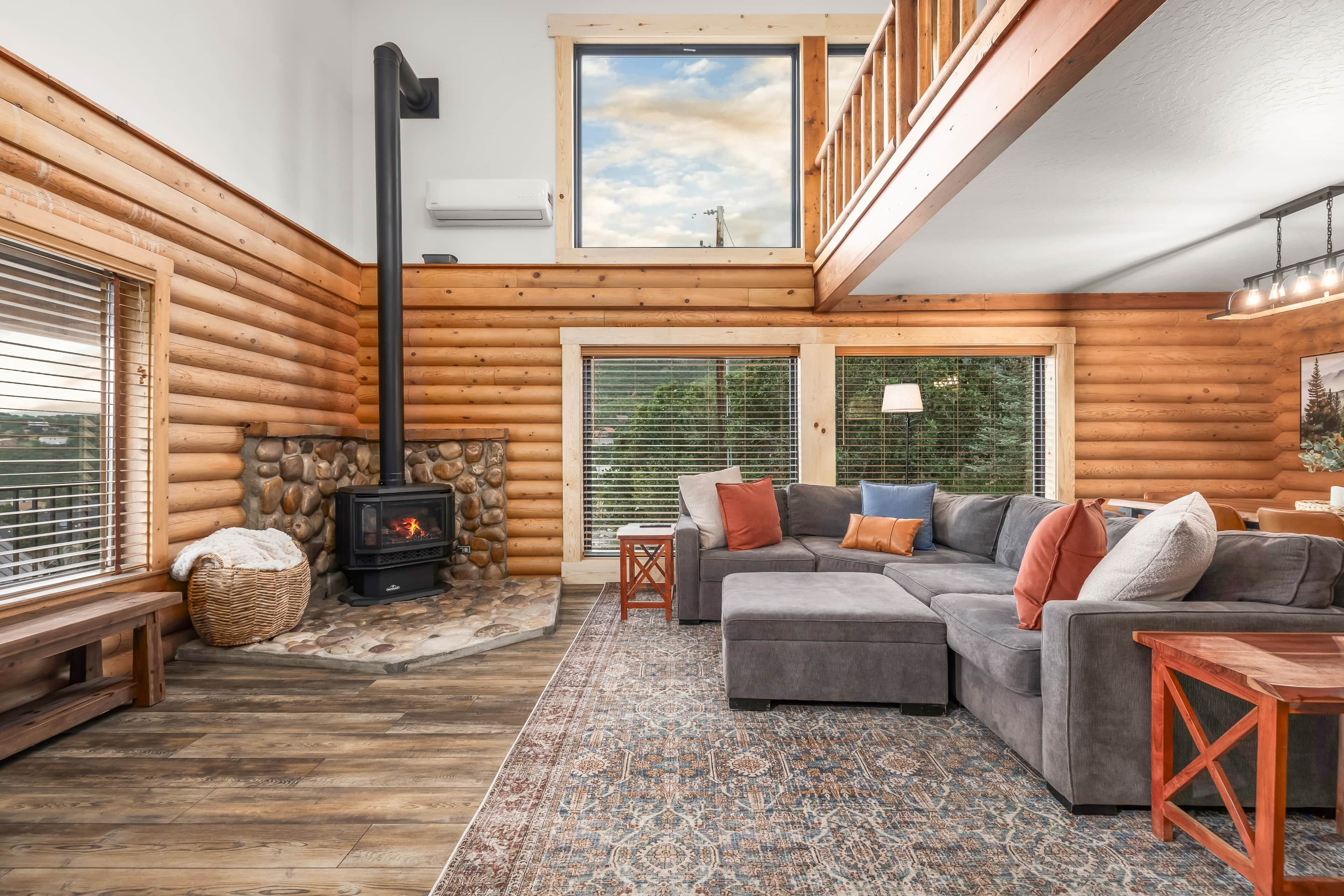 A log cabin interior with a stone fireplace, a sectional sofa, and a large window.