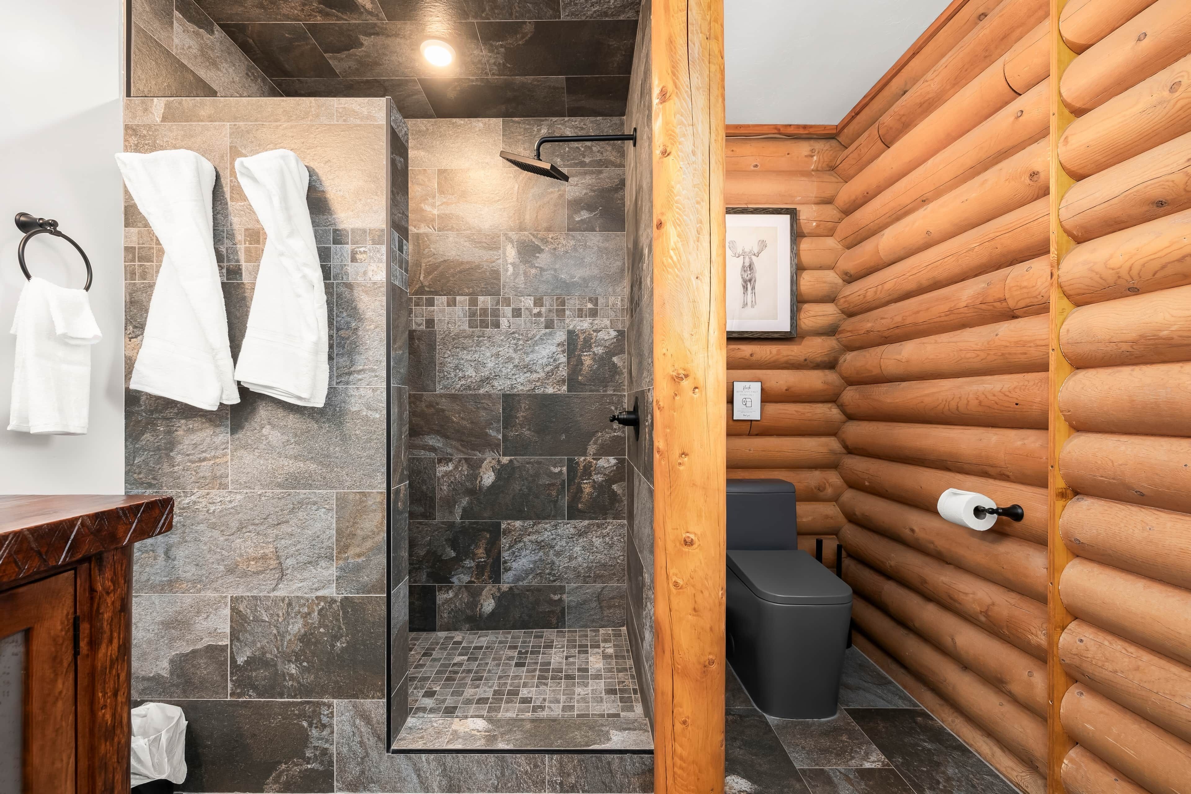 A bathroom with stone tiles, a shower, a wooden log wall, a toilet, and a sink with a towel rack.