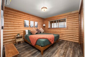 Ein Schlafzimmer in einer Blockhütte mit einem Bett, einem Nachttisch, einem Fenster mit Jalousien und drei gerahmten Bildern an der Wand.