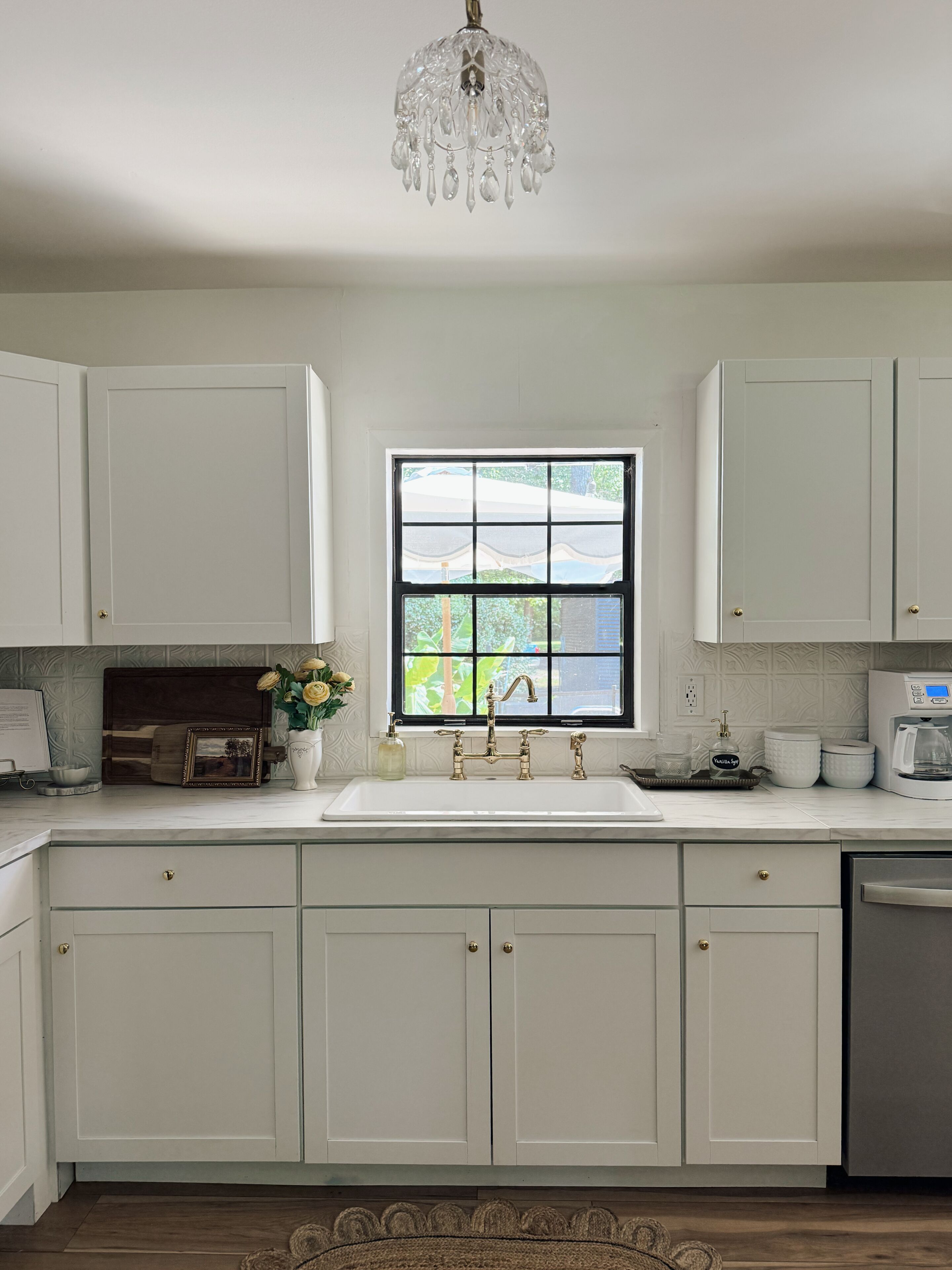 A kitchen with white cabinets, a window with a view, a chandelier, and a coffee maker.