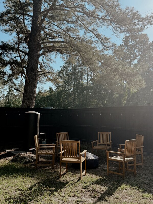 An outdoor seating area with wooden chairs and a table, surrounded by trees and a black wall.