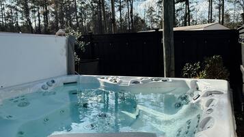 A hot tub with a control panel, surrounded by a wooden deck and a fence, with trees in the background.
