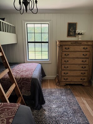 A bedroom with a bunk bed, a dresser, a window with a view of greenery, and a chandelier.