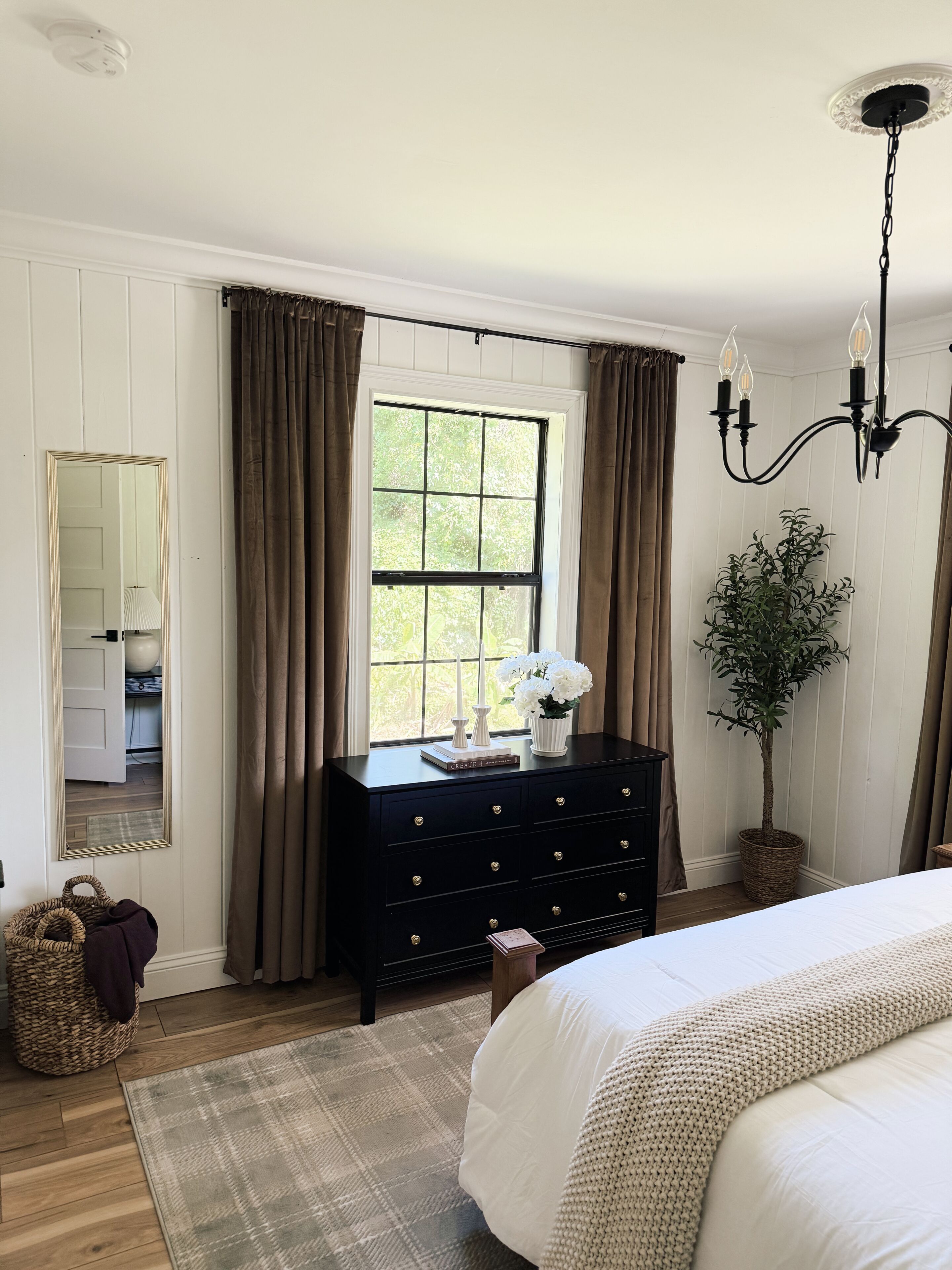 A bedroom with a bed, a dresser, a mirror, a window with curtains, a chandelier, and a potted plant.