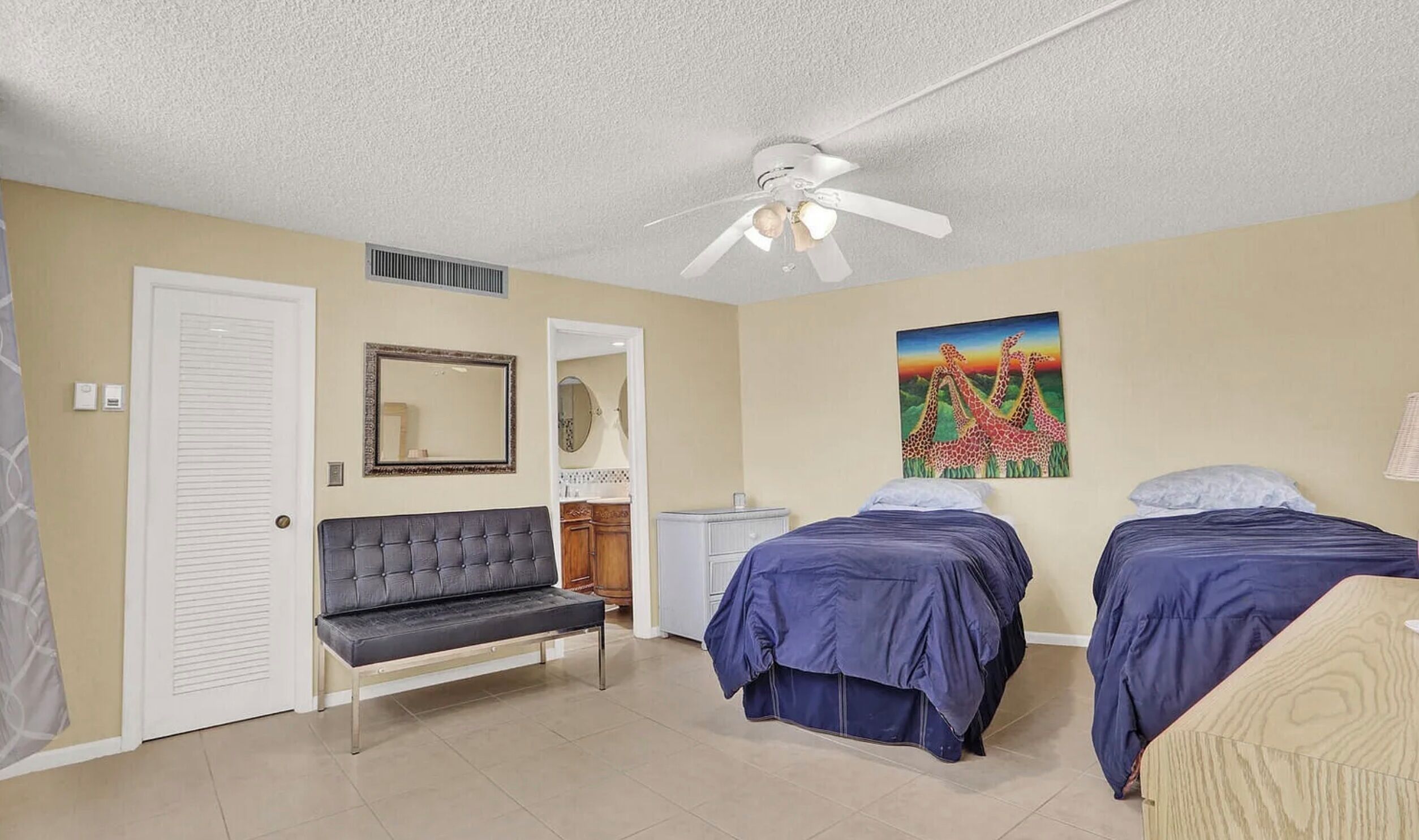 A bedroom with two beds, a bench, a ceiling fan, a painting, and a door.