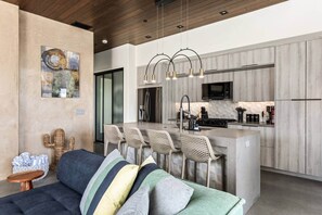 A modern kitchen with a central island, bar stools, and a dining area with a sofa and cushions.
