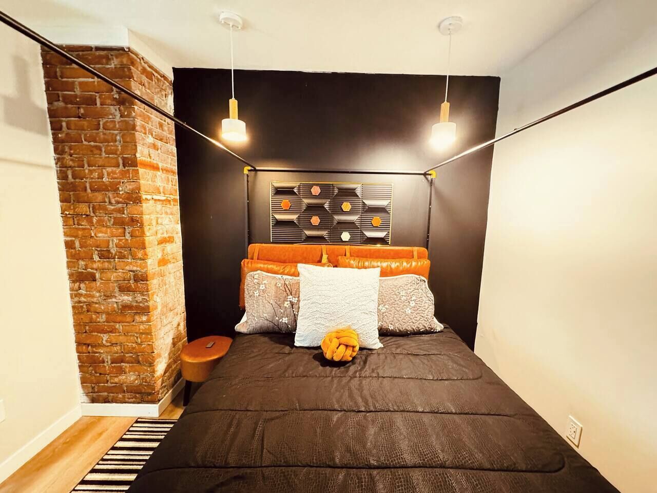 A bedroom with a brick wall, a bed with orange and white bedding, and a geometric wall decoration.