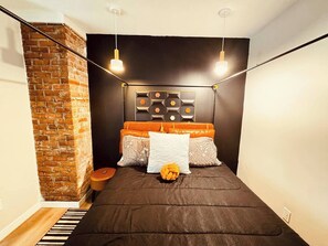 A bedroom with a brick wall, a bed with orange and white bedding, and a geometric wall decoration.