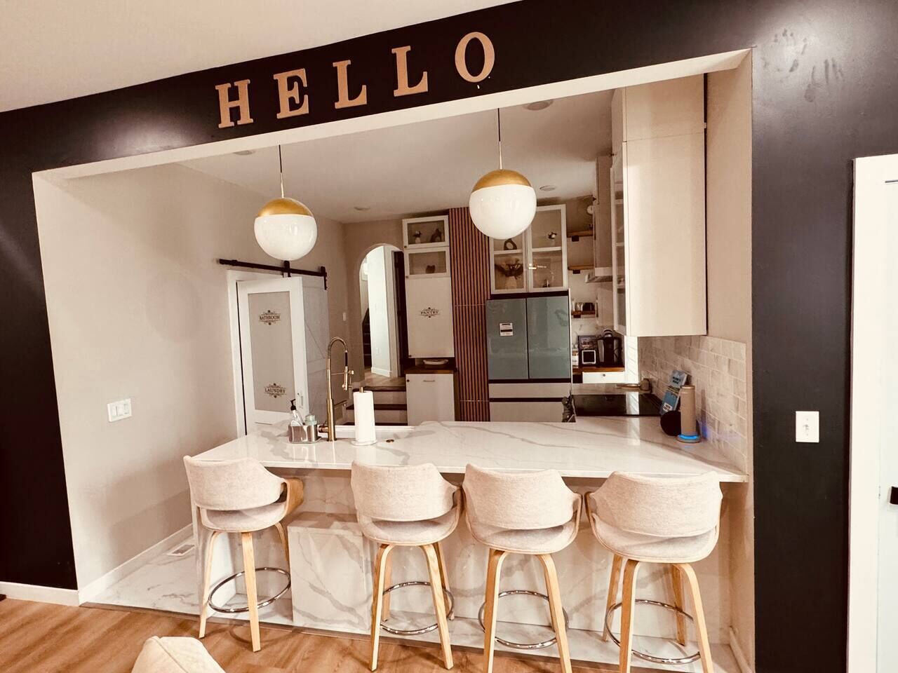 Une cuisine moderne dotée d’un îlot en marbre, de tabourets de bar, de luminaires suspendus et d’une enseigne « BONJOUR » au-dessus de l’entrée.
