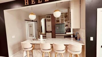 Une cuisine moderne dotée d’un îlot en marbre, de tabourets de bar, de luminaires suspendus et d’une enseigne « BONJOUR » au-dessus de l’entrée.