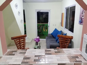 Una sala de estar con un sofá, dos sillas de madera, una mesita con un florero y un ventanal corredizo que da a un balcón.