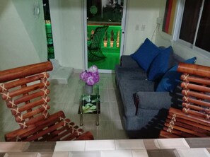 Una sala de estar con un sofá gris, cojines azules, una mesa de centro de vidrio con una flor morada y sillas de madera con diseños en forma de X.