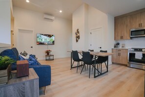 Uma sala de estar moderna com um sofá azul, uma mesa de jantar com cadeiras, uma TV de tela plana na parede e uma área de cozinha com armários de madeira e micro-ondas.