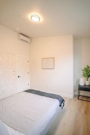 A bedroom with a bed, an air conditioning unit, a door, a framed picture on the wall, a bedside table with a plant, and a string of lights.