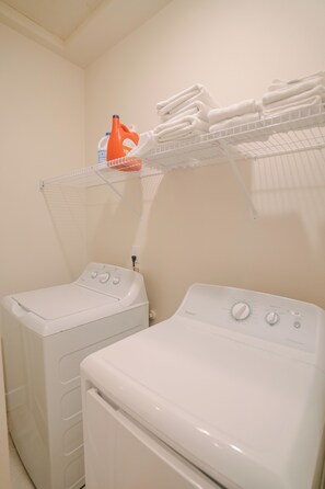A laundry area with two white front-loading washing machines and a shelf above holding folded towels and detergent bottles. - Luxury Home in Heart of Vancouver / Quiet & Cozy (Vancouver)
