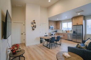 A modern kitchen with stainless steel appliances, a dining area with a wooden table and chairs, and a living room with a sofa and a coffee table.
