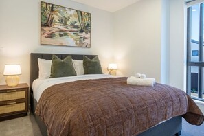 Quarto de hotel com uma cama grande, duas luminárias de cabeceira, um quadro acima da cama e uma janela com vista.