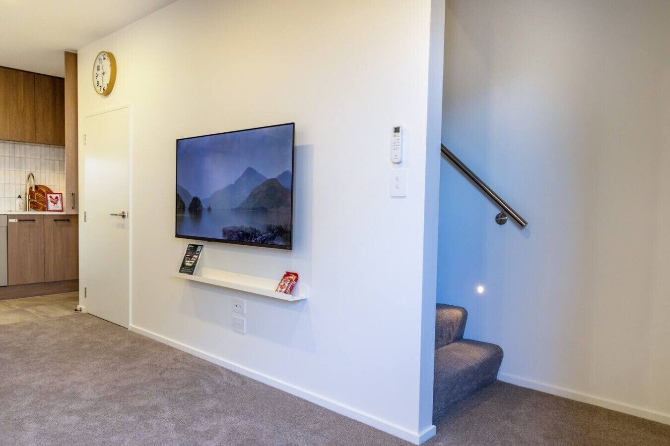 Una sala de estar con un televisor montado en la pared, un estante con objetos, un reloj y una escalera.