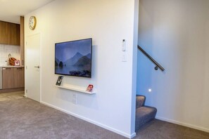 A living room with a mounted TV, a small shelf with items, a clock, and a staircase. - City Fringe 2BR 2BA Townhouse | Parking + EV (Christchurch)