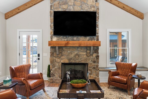 A living room with a stone fireplace, leather chairs, a coffee table, and a flat-screen TV mounted on the wall.