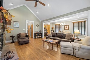 Ein geräumiges Wohnzimmer mit einem braunen Ledersofa, einem beigefarbenen Sessel und einem Holztisch. Eine Deckenventilator und eingelassene Beleuchtung sind sichtbar.