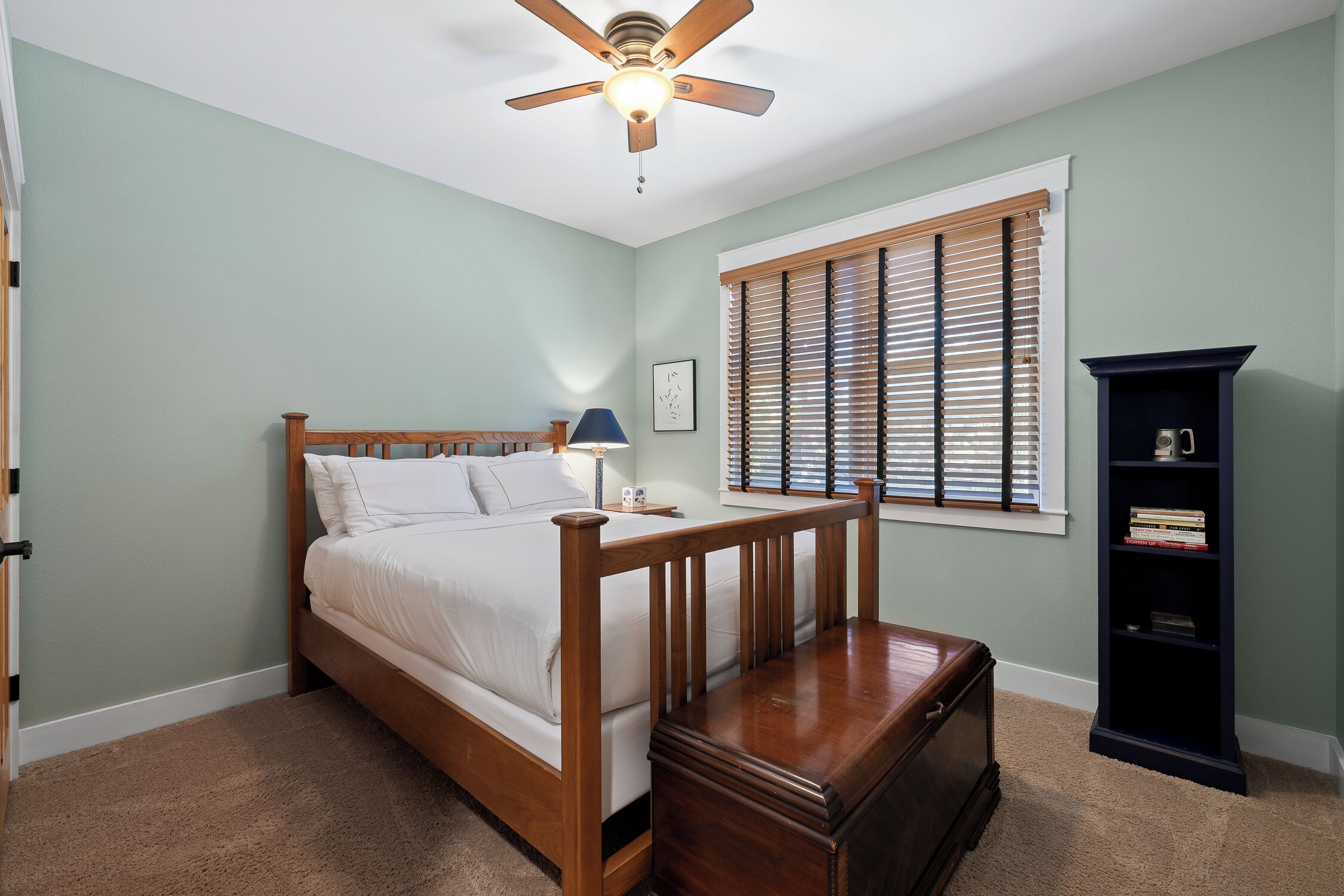 A bedroom with a wooden bed, a nightstand, a lamp, a bookshelf, and a window with blinds.