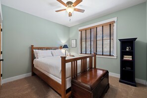 Une chambre à coucher avec un lit en bois, une table de chevet, une lampe, une bibliothèque et une fenêtre avec des stores.