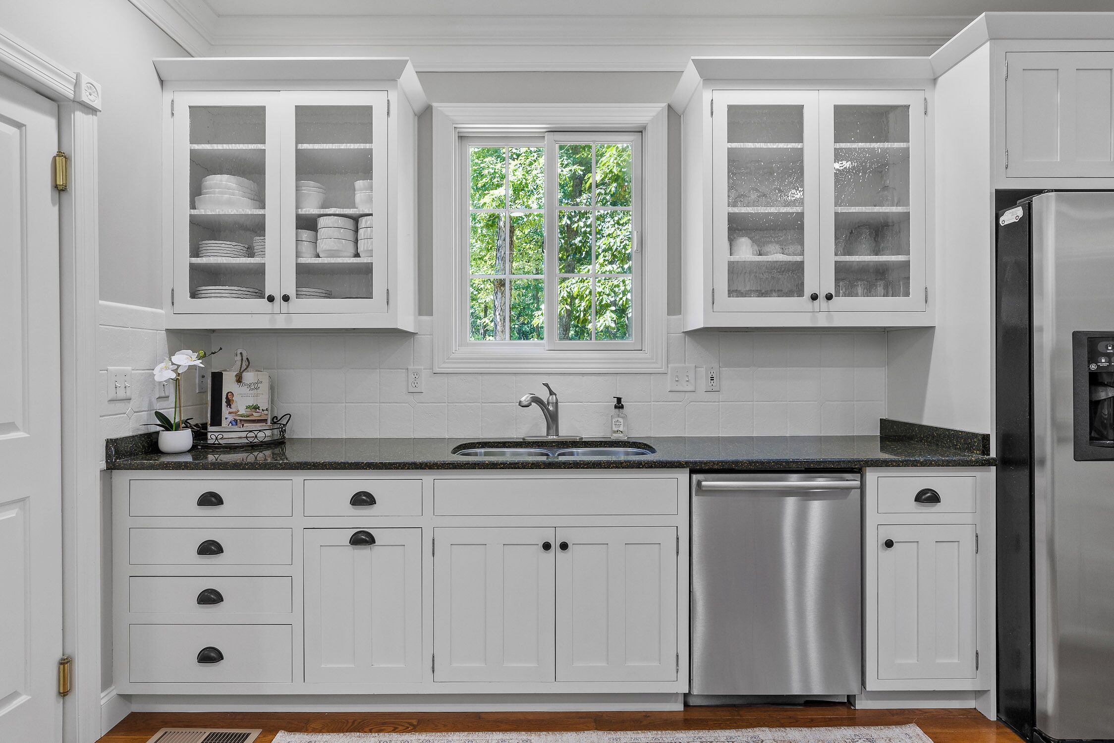 Cocina con armarios blancos, lavavajillas de acero inoxidable y ventana con vista a follaje.
