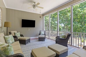 Ein Wintergarten mit Deckenventilator, Flachbildfernseher, Korbmöbeln und Blick auf Bäume durch große Fenster.