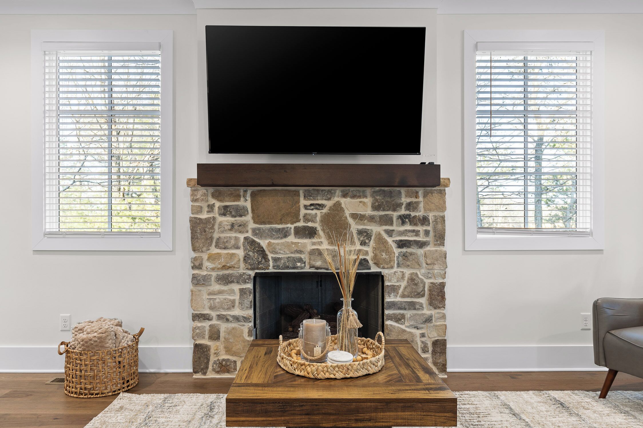 A living room with a stone fireplace, a flat-screen TV mounted above it, and two windows with blinds.
