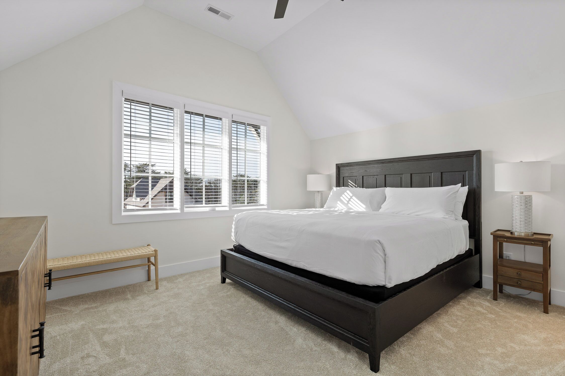 A bedroom with a dark wooden bed, white bedding, a bench, a bedside table with a lamp, and a window with blinds.