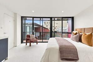 A modern bedroom with a large bed, a chair, and a view through a sliding glass door.