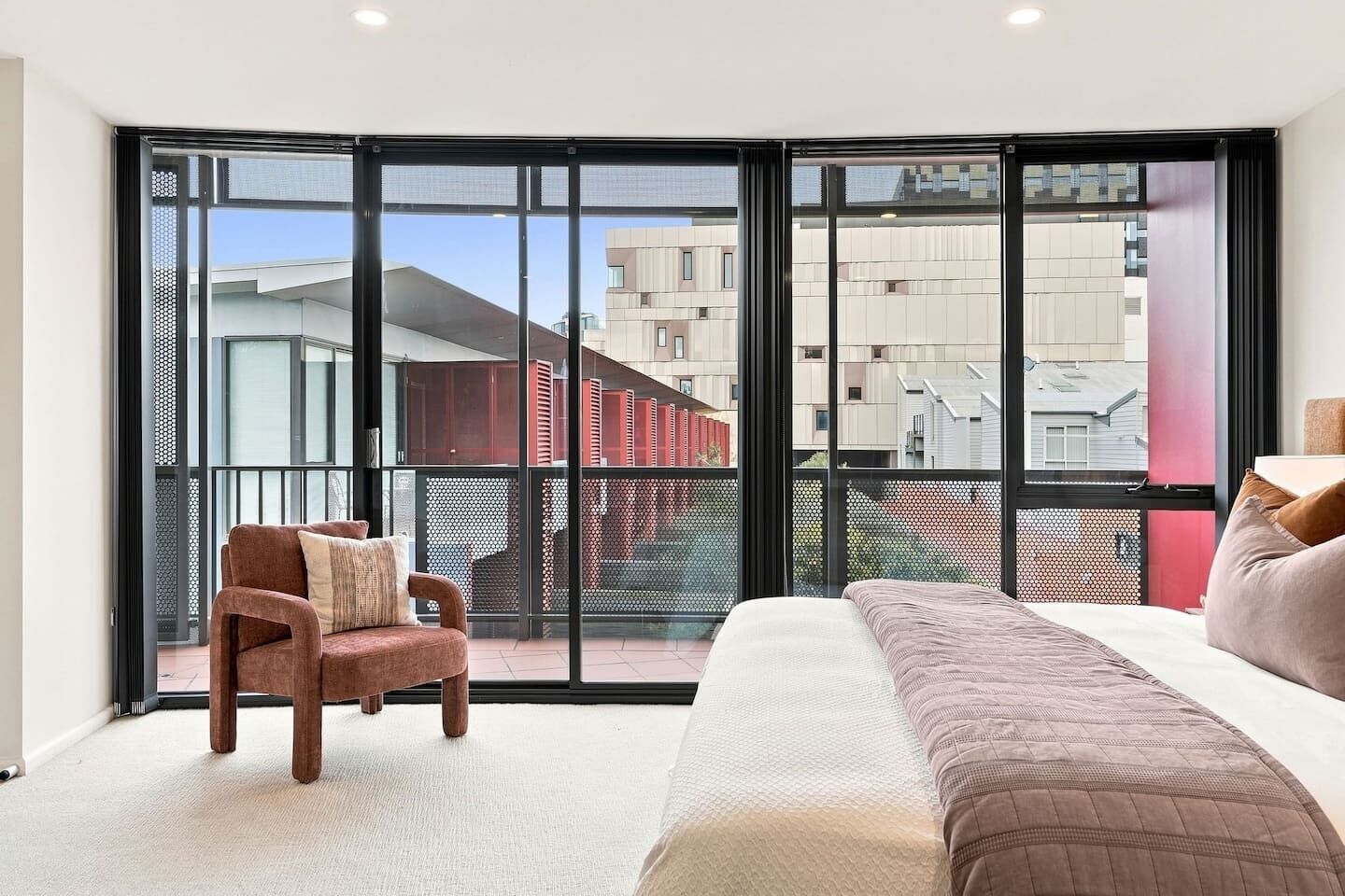 Une chambre avec une grande fenêtre, un lit, une chaise et une vue sur les bâtiments à l’extérieur.