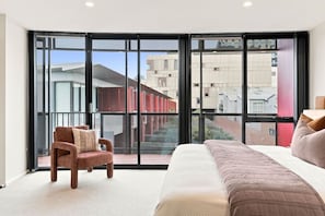 A bedroom with a large window, a bed, a chair, and a view of buildings outside.