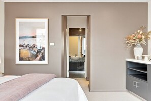 A hotel room with a bed, a framed picture on the wall, a bathroom with a sink, and a vase with flowers on the side.