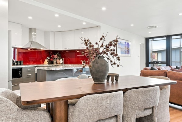 A modern kitchen with white cabinets, a red backsplash, and a central island. There is a dining table with chairs in the foreground.
