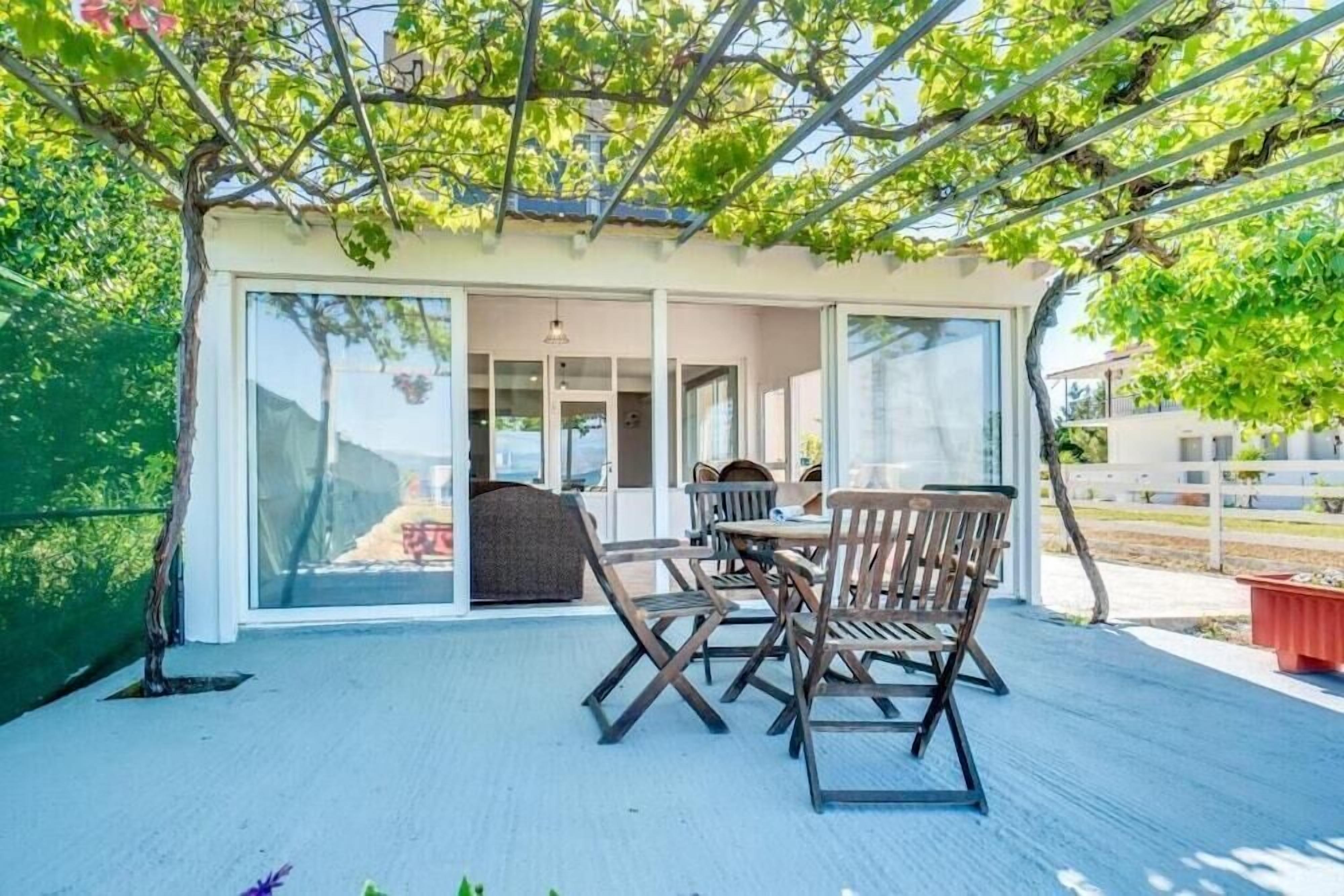 Eine Terrasse im Freien mit Tisch und Stühlen, ein überdachter Bereich mit einer Pergola und Blick ins Haus.