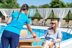Un área junto a la piscina con una mesa y sillas, una persona con una camisa azul y otra sentada a la orilla.