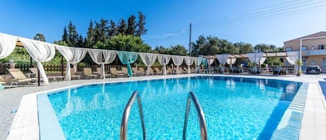 A swimming pool with clear blue water, surrounded by lounge chairs and umbrellas, with a building in the background.
