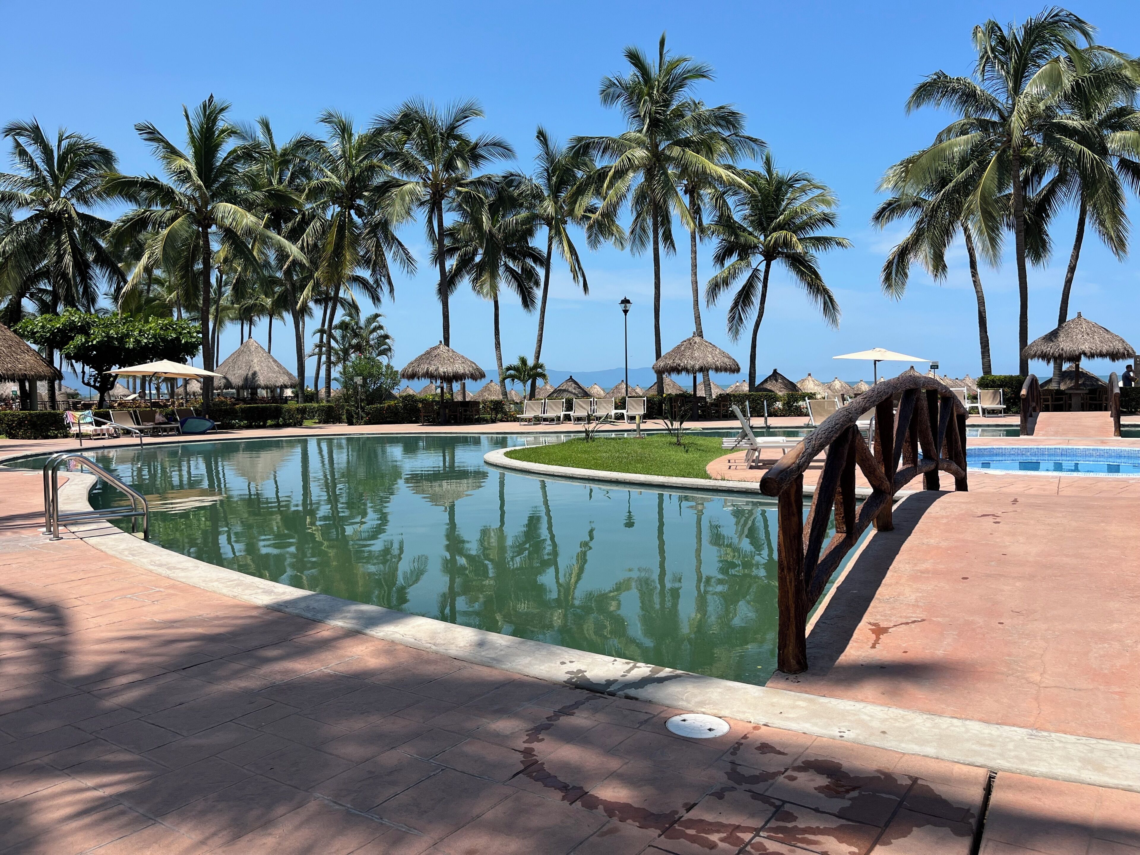 Un área de piscina en un resort con palmeras, sillones de descanso y sombrillas de paja.